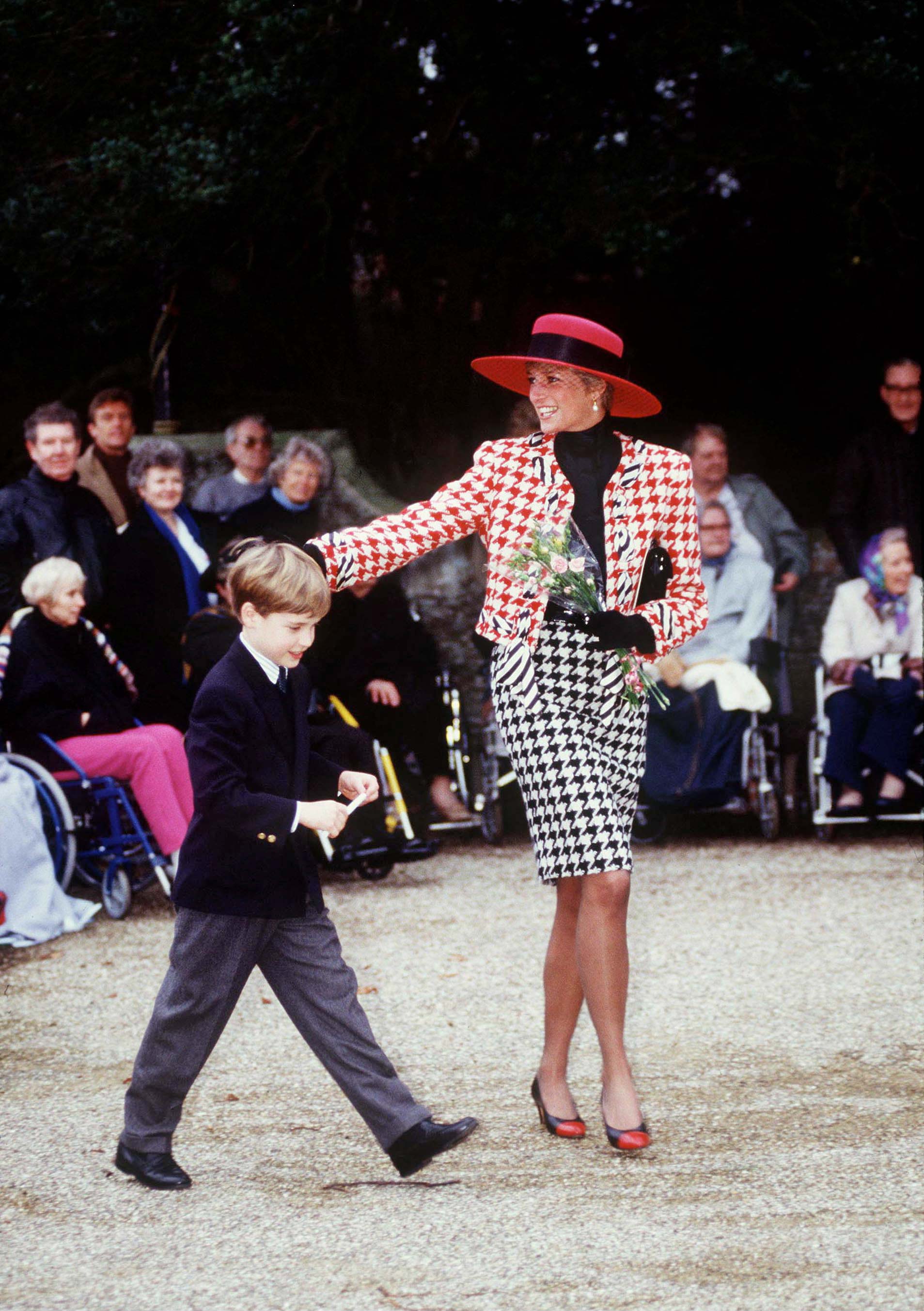 Diana de Gales con uno de sus hijos. Lleva un traje de chaqueta y falda con estampado de color negro y rojo.