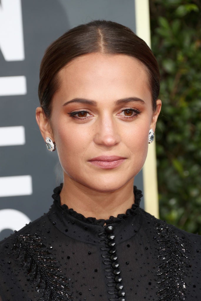 Alicia Vikander con moño pulido y raya en el medio y un discreto maquillaje con sombras marrones, eyeliner negro y labios nude.