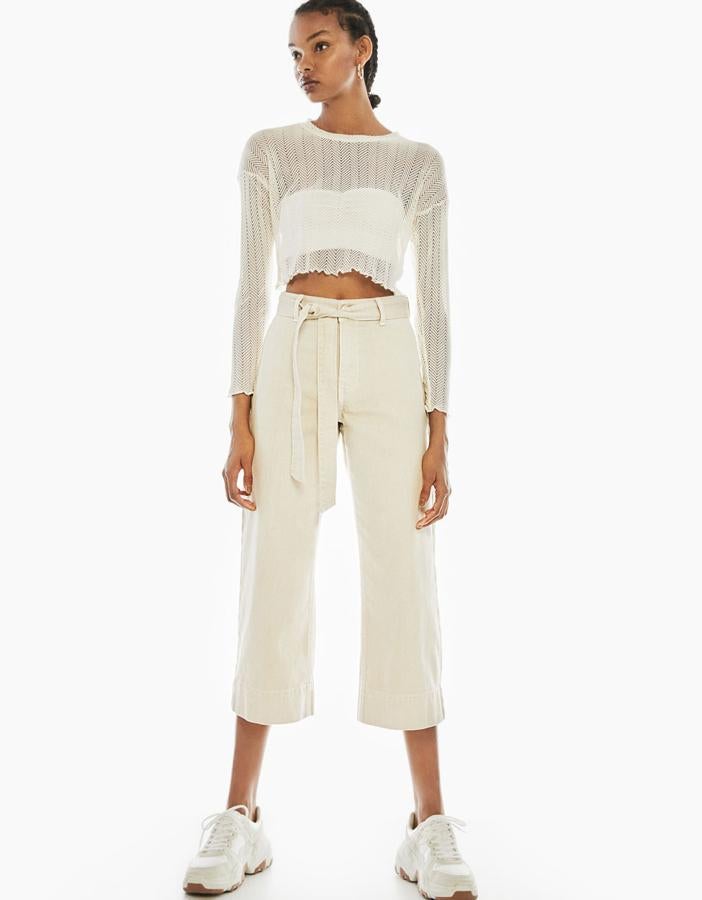 Pantalón denim con tiro alto, pierna ancha y a la altura de los tobillos, con un cinturón en el mismo tejido (25,99 euros). Los lleva con un jersey de punto calado con manga corta y cuello redondo (9,99 euros) y unas deportivas combinadas con la suela XL (35,99 euros).