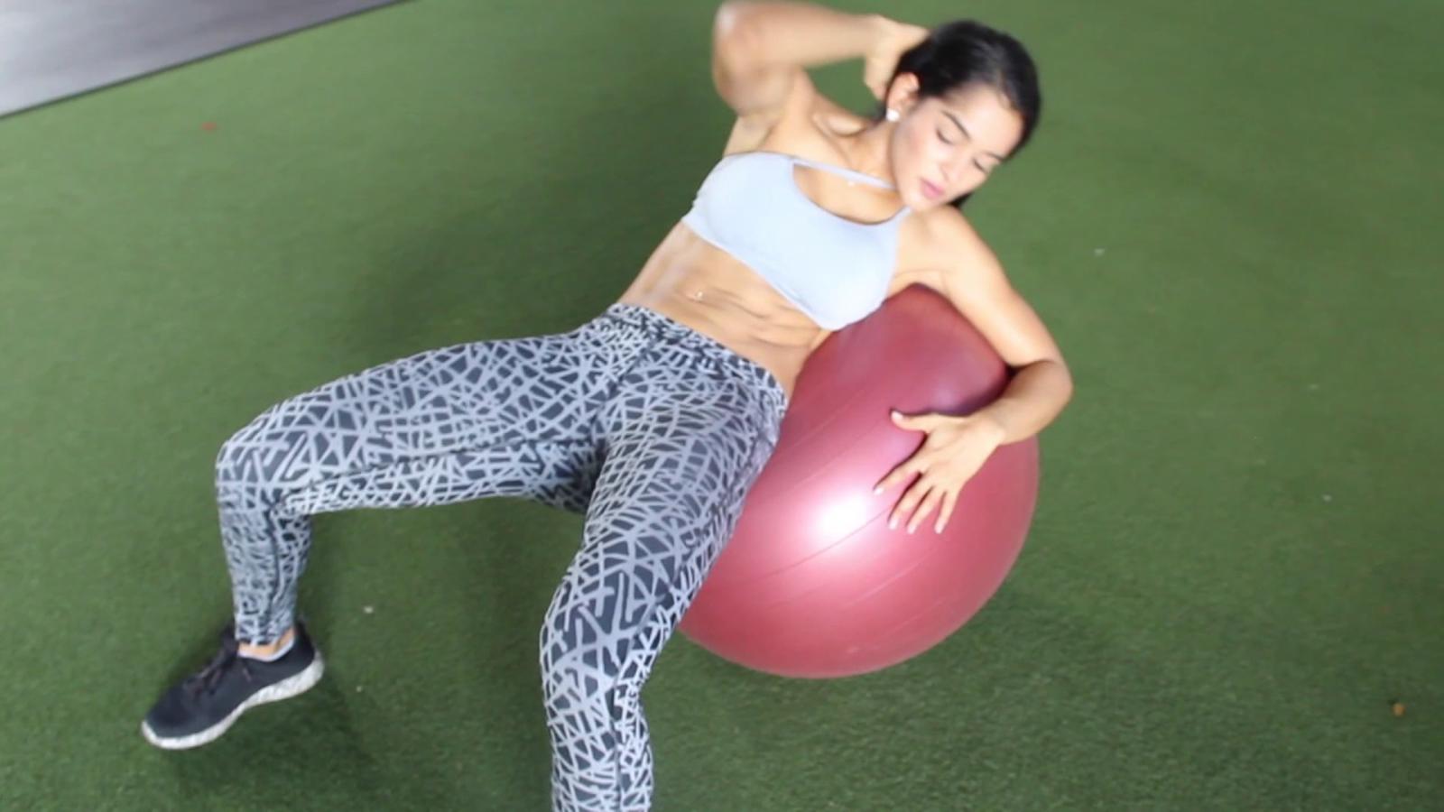 Siéntate sobre la pelota y túmbate hacia atrás y trae tu codo derecho a tu rodilla izquierda como si fuese unos abdominales de bicicleta. Alterna el ejercicio. Repite 4 series de 12 repeticiones.