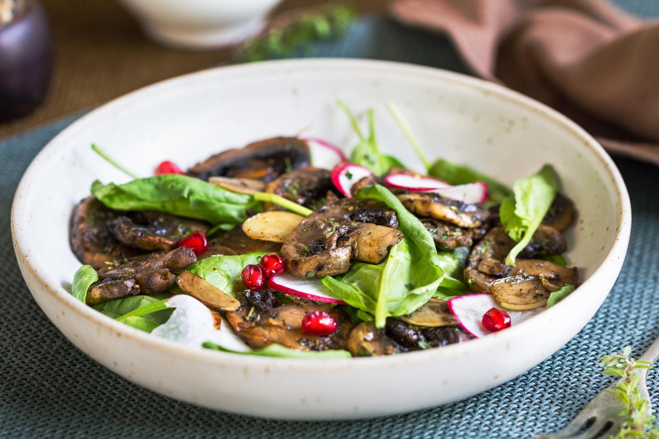 Sencillo y delicioso. Pon una cama de espinacas aliñada con aceite y sal en un plato. A continuación, lamina los champiñones y pon unos ajos a freír en la sartén con algo de aceite. Cuando estén dorados, añade los champiñones, salpimentados y cocínalos hasta el punto que más te guste. Viértelos sobre la cama de lechuga decora con rabanitos y granada.
