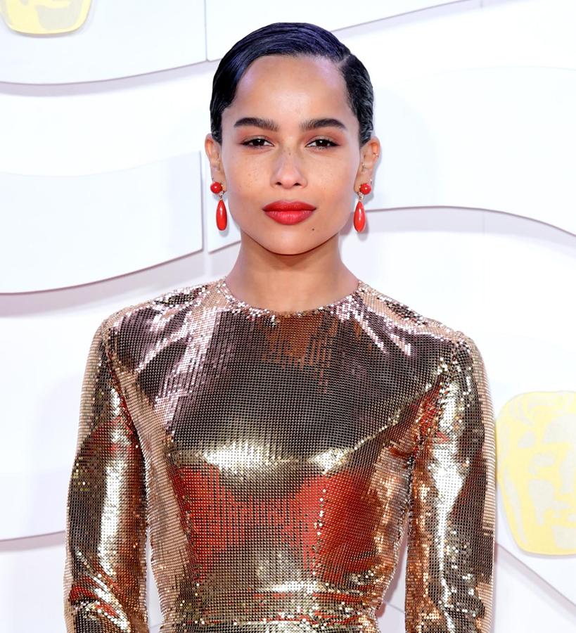 Zoe está de lo más favorecida con su maquillaje de tonos cálidos, que combina labios rojos, con un toque anaranjado a juego con sus pendientes, con sombras ligeramente rojizas. Completa su look pelo engominado con raya lateral.