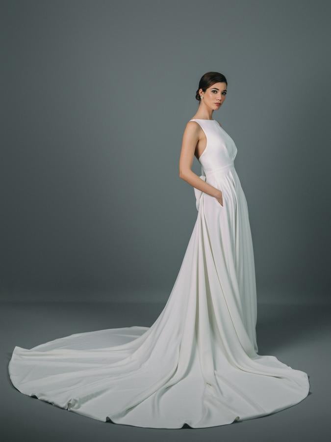 Vestido de novia confeccionado en crepé con cuerpo fruncido y espalda descubierta. Detalle de gran lazada en la espalda. Corte en la cintura y falda de capa con bolsillos y cola.