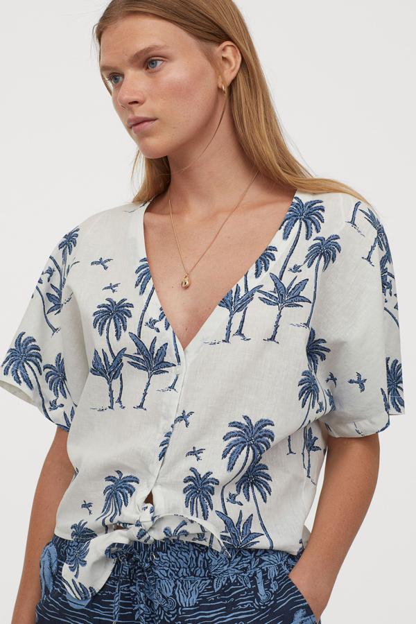De manga corta y escote de pico pronunciado, esta blusa de tejido vaporoso está confeccionada en lino y algodón. Con bajo anudado, tiene un precio de 19,99 euros y estampado de palmeras en tonos azules.