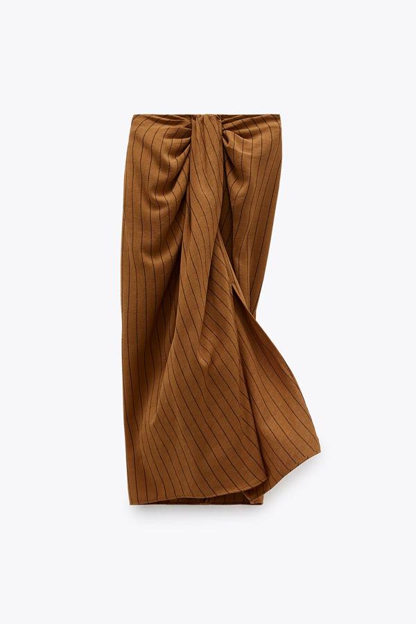 Esta falda pareo de tiro alto con detalle de nudo y pliegues en la parte delantera cuenta con abertura frontal. En color tostado, tiene un precio de 29,95 euros.