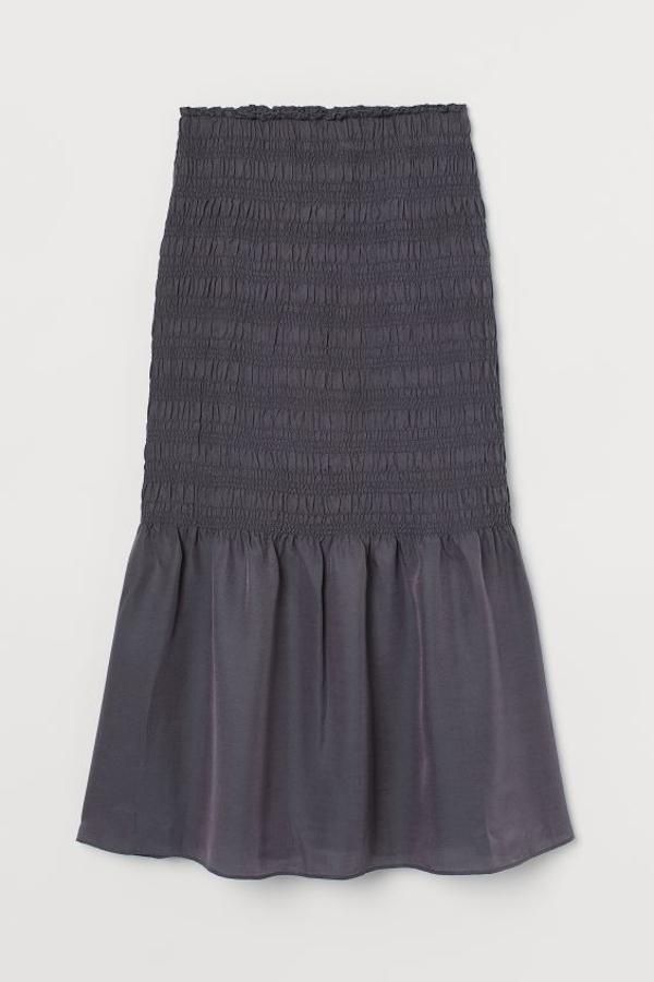 Esta falda midi de H&M en gris oscuro está confeccionada en sarga ligera fluida de mezcla de lyocell sostenible y seda, que le permite cambiar de color según la luz. Con cintura ancha con elástico, tejido fruncido hasta la rodilla y sección acampanada tras ella, tiene un precio de 129 euros y está disponible entre las tallas 34 y 46, y en beige oscuro.