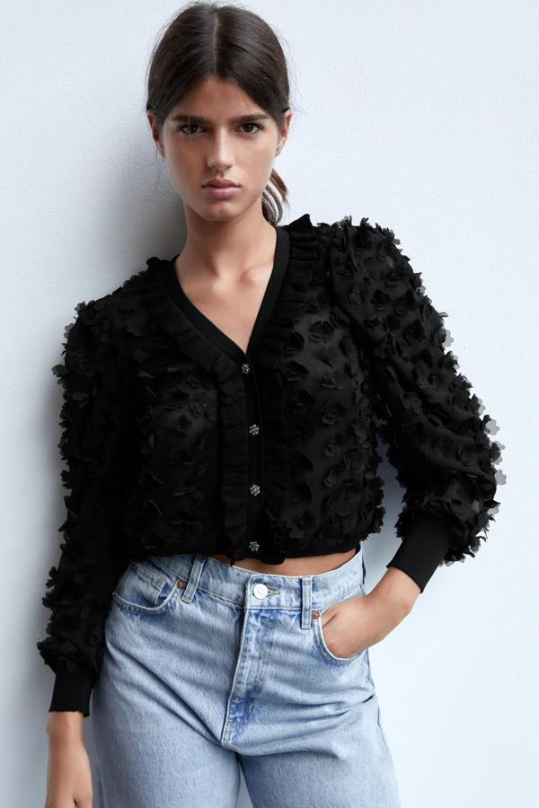 Esta chaqueta de punto con aplicación de tejido en forma de flor cuenta con cierre frontal mediante botones joya. En color negro, es de Zara y tiene un precio de 29,95 euros.