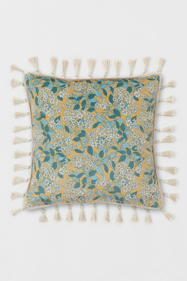 Esta funda de cojín en terciopelo de color amarillo mostaza con estampado floral en tonos azules cuenta con ribete de borlas y cremallera oculta. Con una medida de 40x40 centímetros, tiene un precio de 14,99 euros.