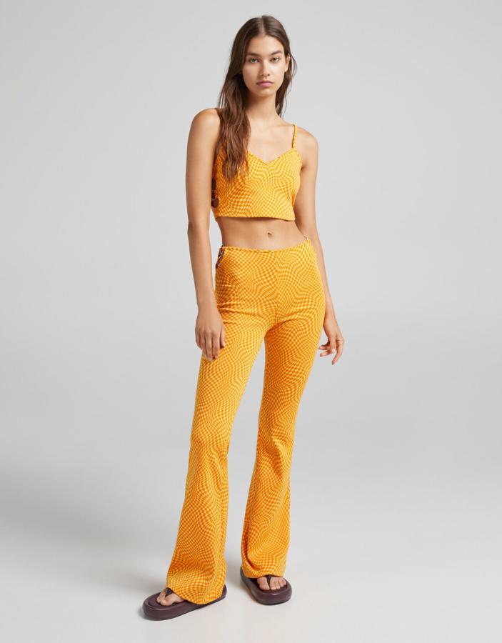 En Bershka han apostado por el estampado psicodélico para este conjunto en amarillo y naranja que está compuesto por un top jacquard con detalle lateral de anillas, que cuesta 12,99 euros, y un pantalón de diseño flare con anillas en la cintura que tiene un precio de 22,99 euros.