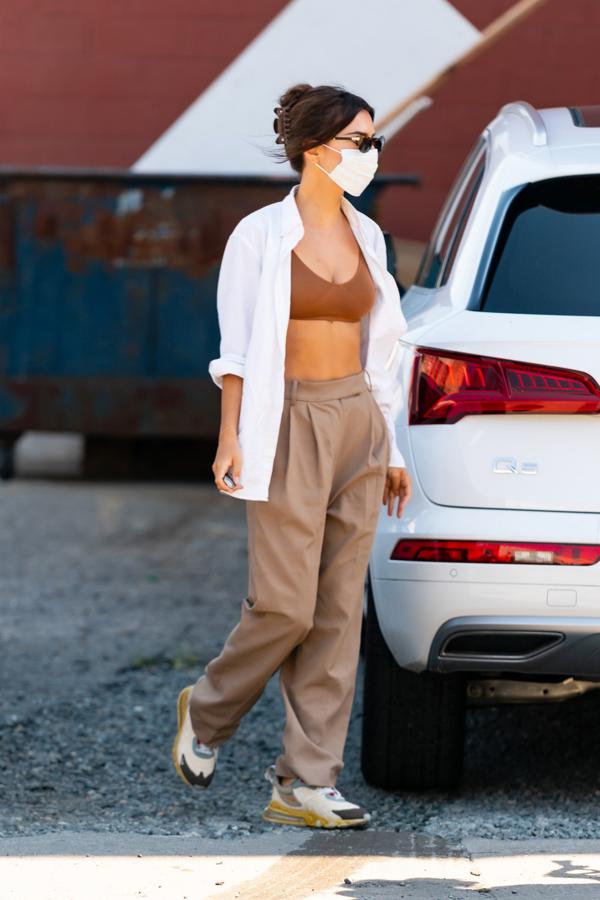 La modelo lleva una camisa blanca oversize abierta para dejar ver el bralette de punto marrón, con pantalón de pinzas beige y deportivas. 