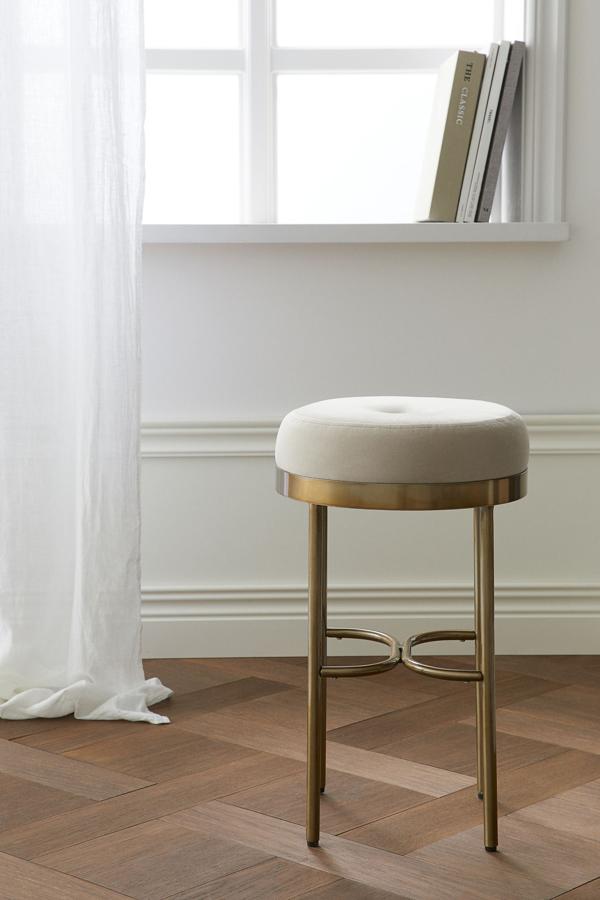 Con cojín acolchado en terciopelo de color beige claro, este taburete redondo de metal dorado tiene una altura de 49 centímetros y un diámetro de 30. Su precio es de 69,99 euros.