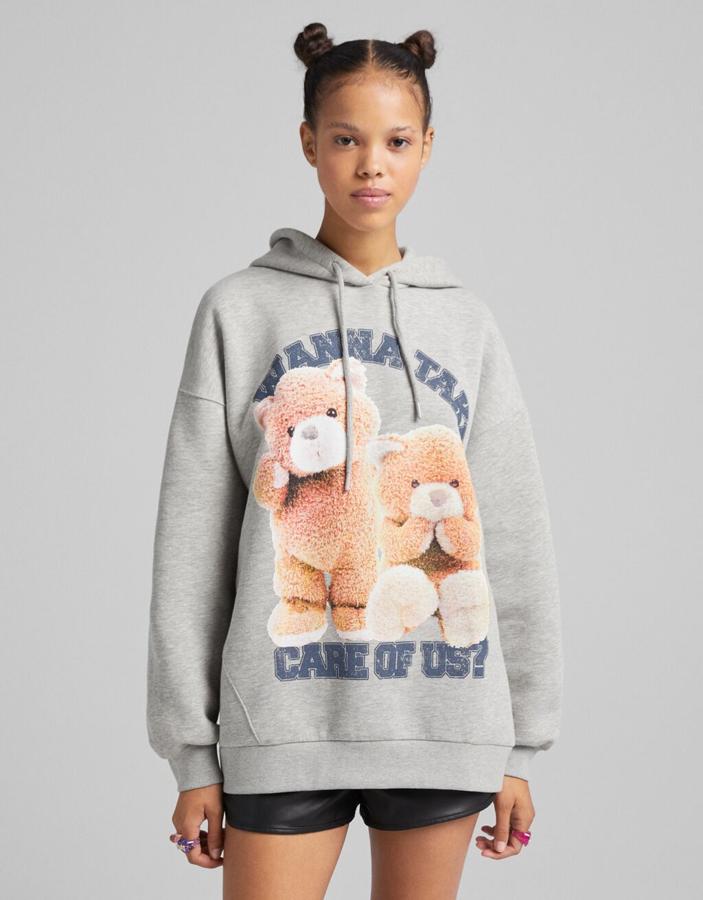 En gris vigoré, con estampación frontal de mensaje en azul y ositos en color anaranjado, esta sudadera de Bershka con capucha ajustable con cordones cuesta 25,99 euros y está disponible entre las tallas XS y L.