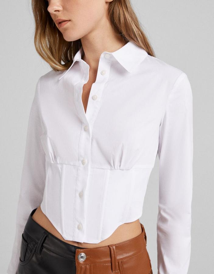 Diseño tipo corsé para esta camisa blanca ajustada de Bershka con cuello de solapa, elástico tipo nido de abeja en la espala y cierre frontal de botones que podemos encontrar entre las tallas XS y L por 19,99 euros. 