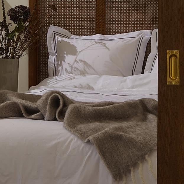 Imagen principal - el juego de funda nórdica en algodón percal en blanco ribeteado cuesta 89.99 euros; la funda de cojín de yute, 39.99 euros; y las mantas de lana, entre 99 y 69.99 euros.