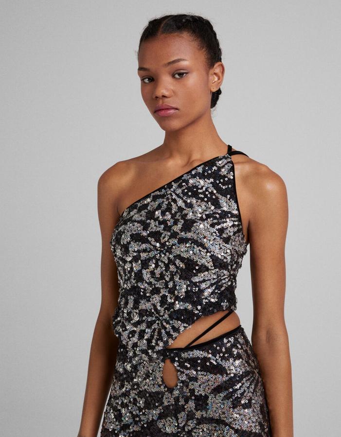 Diseño asimétrico con lentejuelas en plata y negro para este top asimétrico de Bershka con un hombro al aire y tirantes finos en la espalda. Cuesta 19,99 euros. 