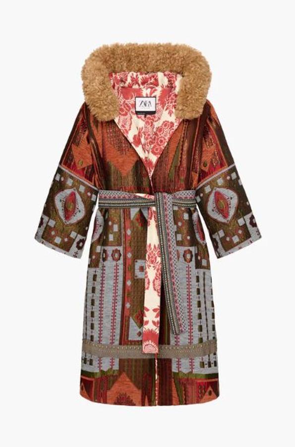 Abrigo largo revestido con jacquard de hilo metalizado y punto de cruz. Lleva una capucha con aplicación efecto pelo extraíble y manga larga. Además, tiene un cinturón lazada en mismo tejido y el detalle de cinta ancha de algodón con tachas piramidales de oro envejecido en el dobladillo (299 euros).