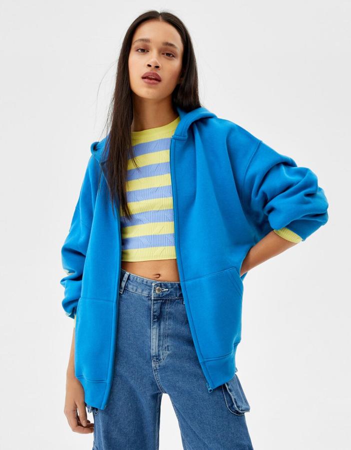 Con print a contraste en la espalda y en una manga, esta chaqueta de diseño oversize y color azul de Bershka con capucha y cierre frontal de cremallera a tono cuesta 25,99 euros.