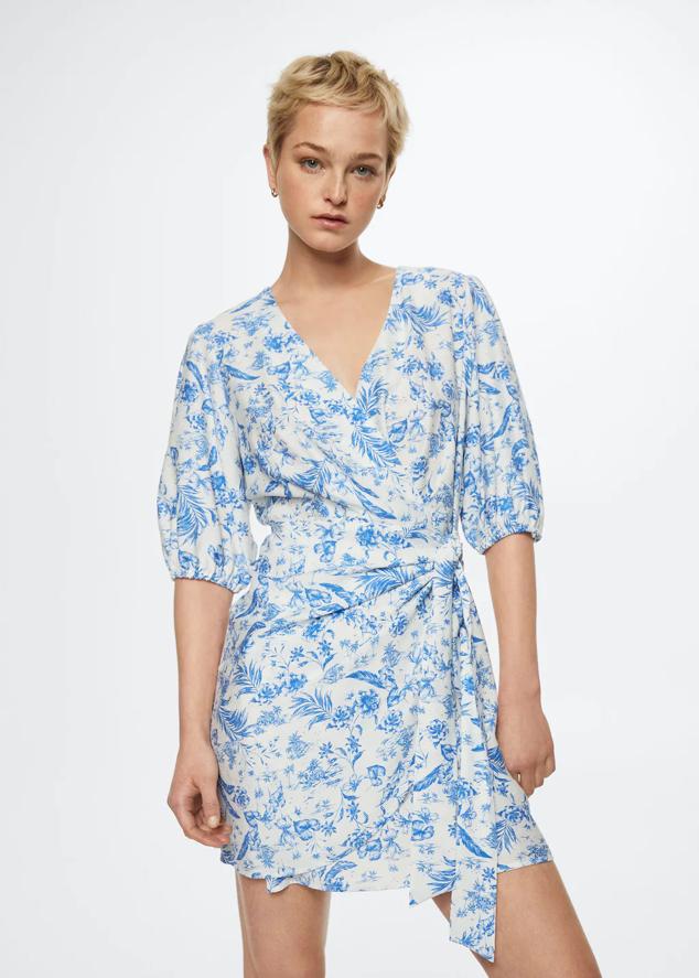 En color blanco con estampado en tonos azules, este vestido corto de Mango confeccionado en tejido fluido con diseño recto y cuello cruzado, viene con mangas cortas abullonadas y tiene cierre de lazo lateral. Tiene un precio de 35,99 euros. 