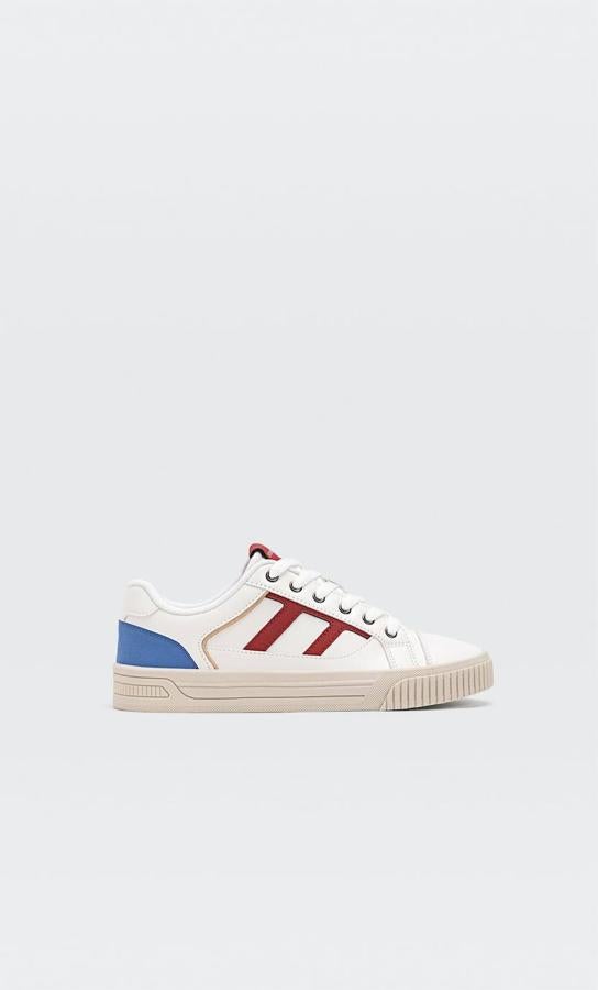Zapatillas deportivas color blanco con bandas laterales en rojo y pieza azul en el talón, suela plana con puntera reforzada y plantilla técnica, de Stradivarius. Tienen un coste de 29,99 euros.