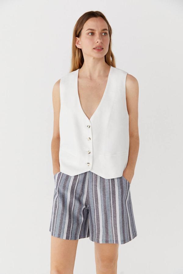 Chaleco de vestir con corte holgado y relajado, escote en uve, maga sisa, bolsillos ribeteados, cierre de botones frontal y bajo asimétrico, color blanco. Puedes comprarlo a 49,99 euros.