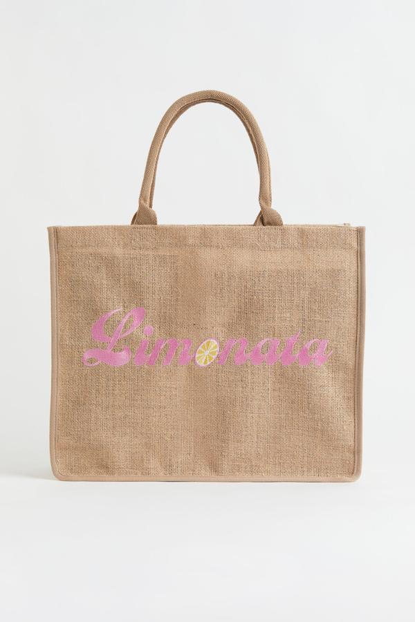 Estilo shopper y de tamaño grande. Está hecha en tejido de yute con motivo estampado delante. Incluso posee dos asas de algodón y ribete en color de contraste. Es de H&M. Precio: 19,99 €.
