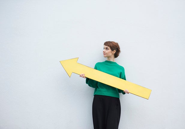 A young student woman stands against a white wall holding a large yellow arrow sign.