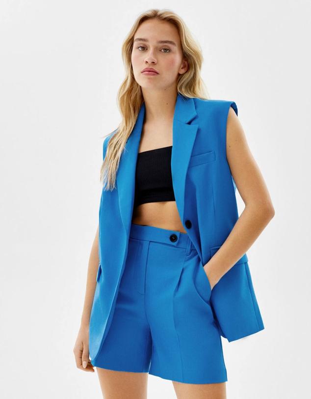 Traje compuesto por chaleco largo de sastre con cuello solapa y cierre de botonees, más unas bermudas de vestir con talle alto, bolsillos y cintura elástica, de Bershka. Está disponible en azul turquesa, beige o negro, a 25,99 euros y 22,99 euros.