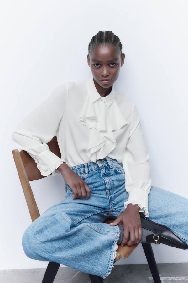 Blusa en tejido fluido con cuello solapa, manga larga terminada en puño, detalles de botones joya y volantes en el pecho, color blanco, de Zara. Cómprala a 25,95 euros.
