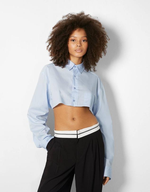 Camisa cropped en tejido de popelín ligero con manga larga, cuello solapa, cierre de botones a tono y bajo sin rematar, en celeste claro, de Bershka. Puedes comprarla a 25,99 euros.