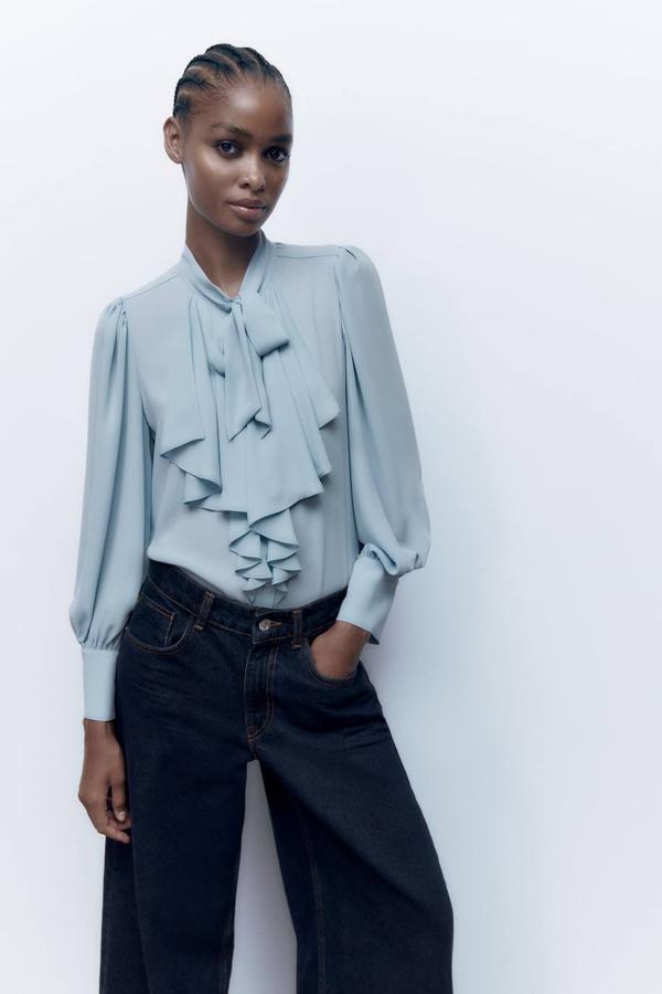 Blusa en tejido ligero con detalle de pechera de volantes, manga larga acabada en puño y lazada al cuello, color azul grisáceo, de Zara. Cómprala por 25,95 euros.
