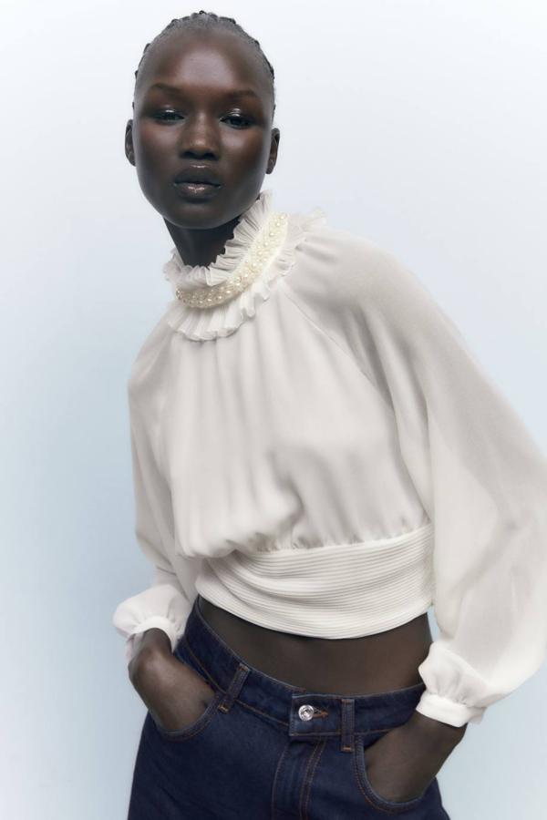 Camisa blanca en tejido fluido vaporoso con bajo plisado, silueta relajada, mangas largas amplias recogidas en puño y cuello subido con volantes y detalles de perlas, de Zara. Cuesta 29,95 euros.
