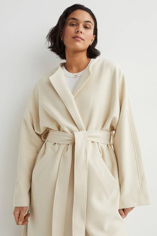 Abrigo de corte recto relaxed fit con cuello redondo, manga larga, bolsillos laterales y cinturón de lazada a combinación, color crema, de H&M. Tiene un coste de 59,99 euros.