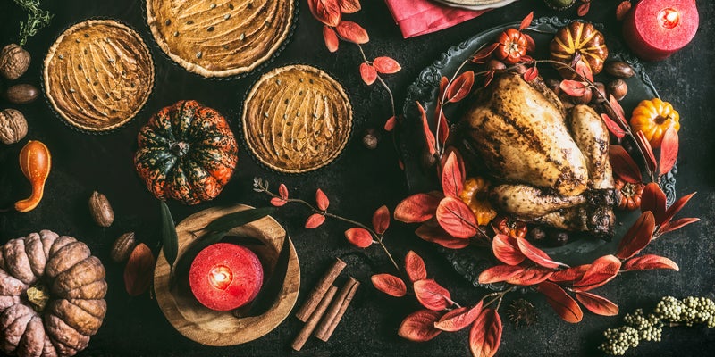 Thanksgiving day dinner table  with roasted whole chicken or little turkey , pumpkin pie on dark rustic background with plates, sauce, cutlery, table setting tags, burning candles, red autumn leaves, nuts and festive decoration , top view, still life, flat lay