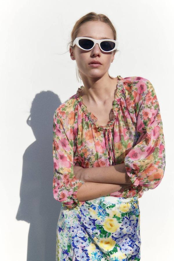 Blusa en tejido vaporoso con hilo metalizado, cuello redondo adornado con volante, lazada y manga por debajo del codo terminada en elástico, en floral print rosa, de Zara. Consíguela a 27,95 euros.