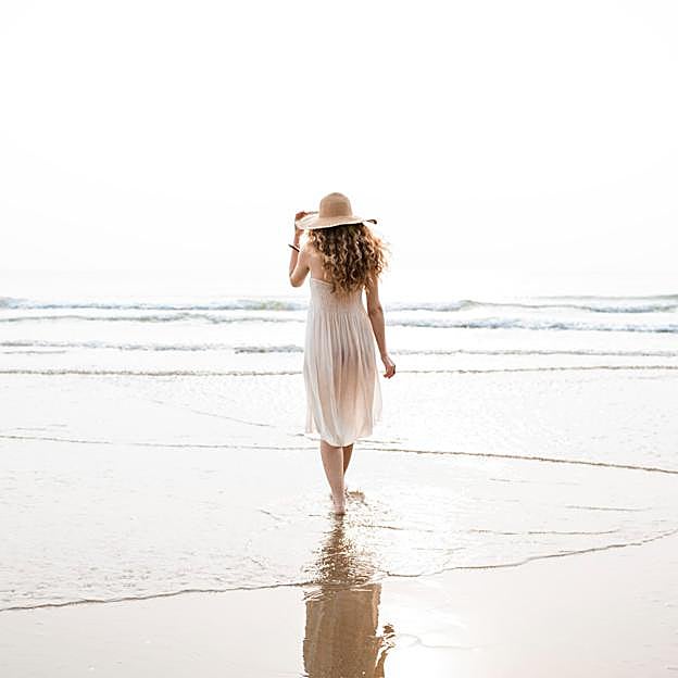 Mujer con vestido blanco y gorro de paja andando por la orilla del mar. 