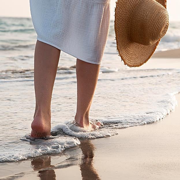 Mujer caminando descalza por la orilla del mar con un gorro de paja en la mano. 