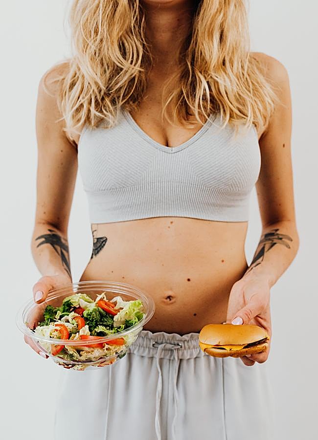 Imagen - Mujer joven decidiendo entre comer una ensalada o una hamburguesa. / Foto de Karolina Grabowska en Pexels.