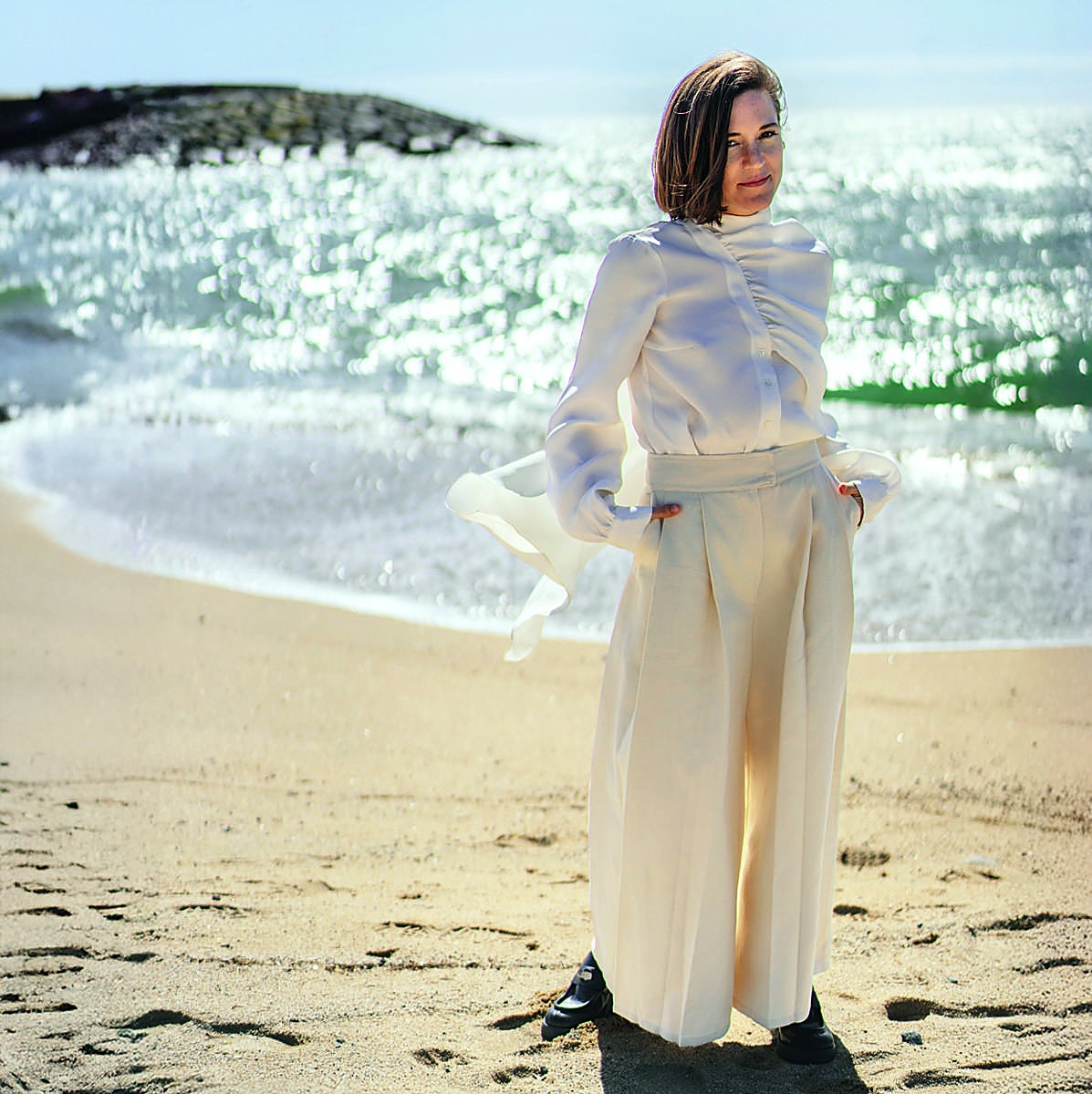 La directora Carla Simón en la playa de El Masnou, con blusa y pantalón de KM by LANGE, sandalias de ABOUT ARIANNE y pendientes de MÖULE..
