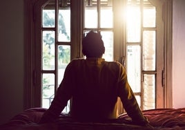 Un joven mira por la ventana sentado en la cama de su habitación.