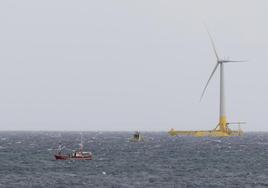 Un barco pesquero junto al prototipo (fondo) de aerogenerador de eólica marina flotante, anclado con cadenas al fondo marino .