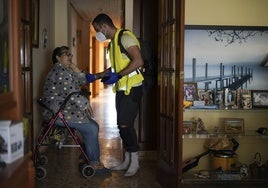 Lola, sentada en su andador en el pasillo de su casa en Catarroja, atendida por un enfermero voluntario.