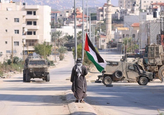 Un hombre lleva una bandera palestina mientras vehículos militares israelíes avanzan por una calle de la ciudad cisjordana de Tulkarem.