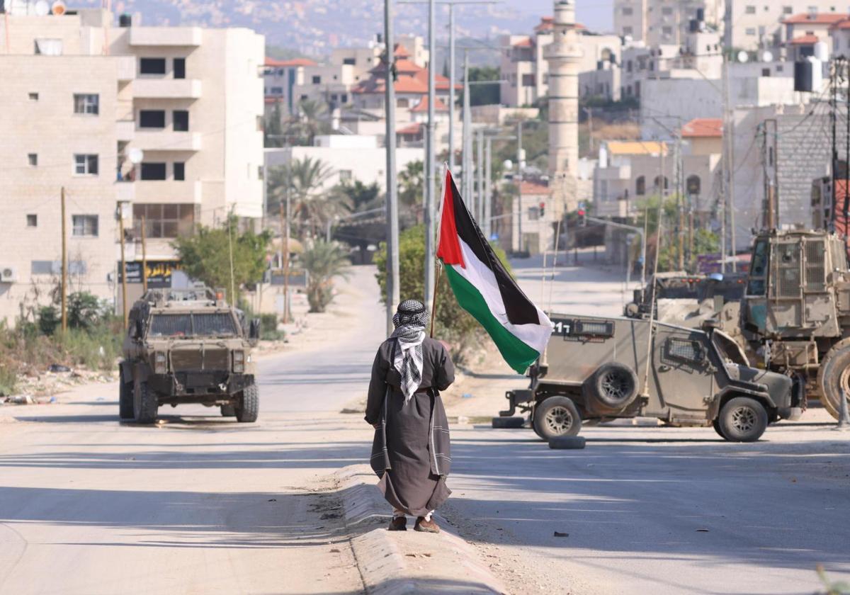 Un hombre lleva una bandera palestina mientras vehículos militares israelíes avanzan por una calle de la ciudad cisjordana de Tulkarem.