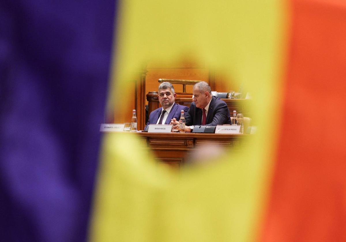 Marcel Ciolacu y un miembro de su equipo ministerial son visibles a través de un recorte de la bandera rumana, que recuerda a la bandera de la Revolución rumana.