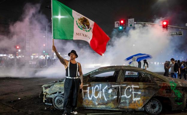 Un joven alza una bandera mexicana junto a un coche quemado.