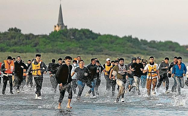 Migrantes corren desde la playa hacia el agua para alcanzar un bote en el que cruzar el canal.