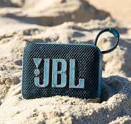 Una colorida naturaleza muerta del altavoz Bluetooth JBL GO 4 sobre una toalla de playa, rodeado de gafas de sol y una bebida, bajo una iluminación soleada y brillante, enfatizando su uso en exteriores.