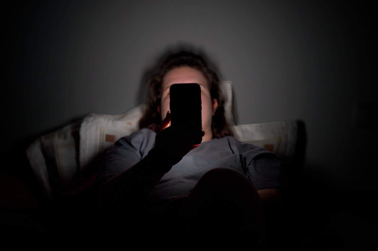 Una joven, tumbada en la cama, mira un teléfono móvil