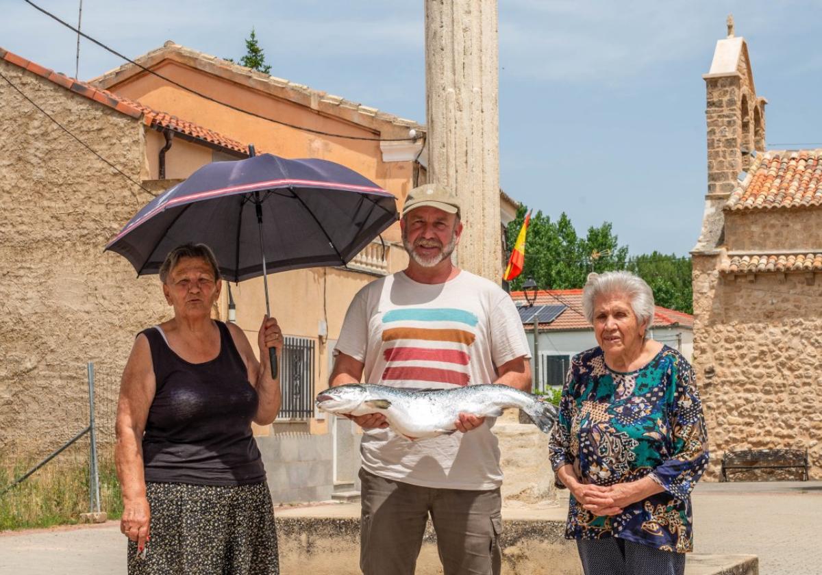 Ángeles und Julia, zwei Einwohnerinnen von Valderrebollo, auf der Plaza Mayor des Dorfes, neben Curro, der einen Lachs hält, wie er ihn ab 2028 vermarkten möchte.