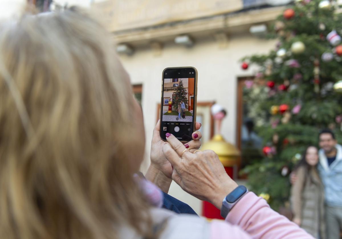 Una mujer hace una foto con el móvil a una pareja junto a un árbol de Navidad.